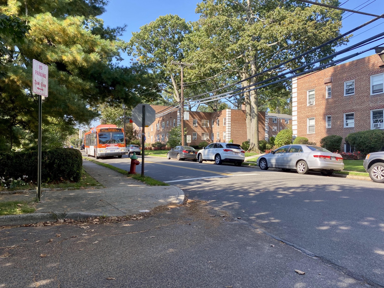 Brick apartment buildings with white-trimmed windows along a street with parked cars and a city bus, in Roslyn Heights