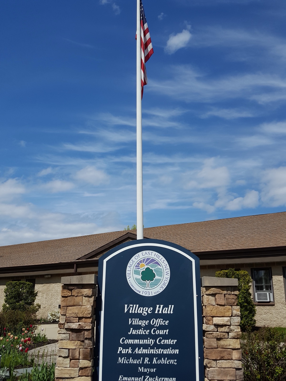 East Hills Village Hall entrance sign with an American flag and a stone-trimmed brick building