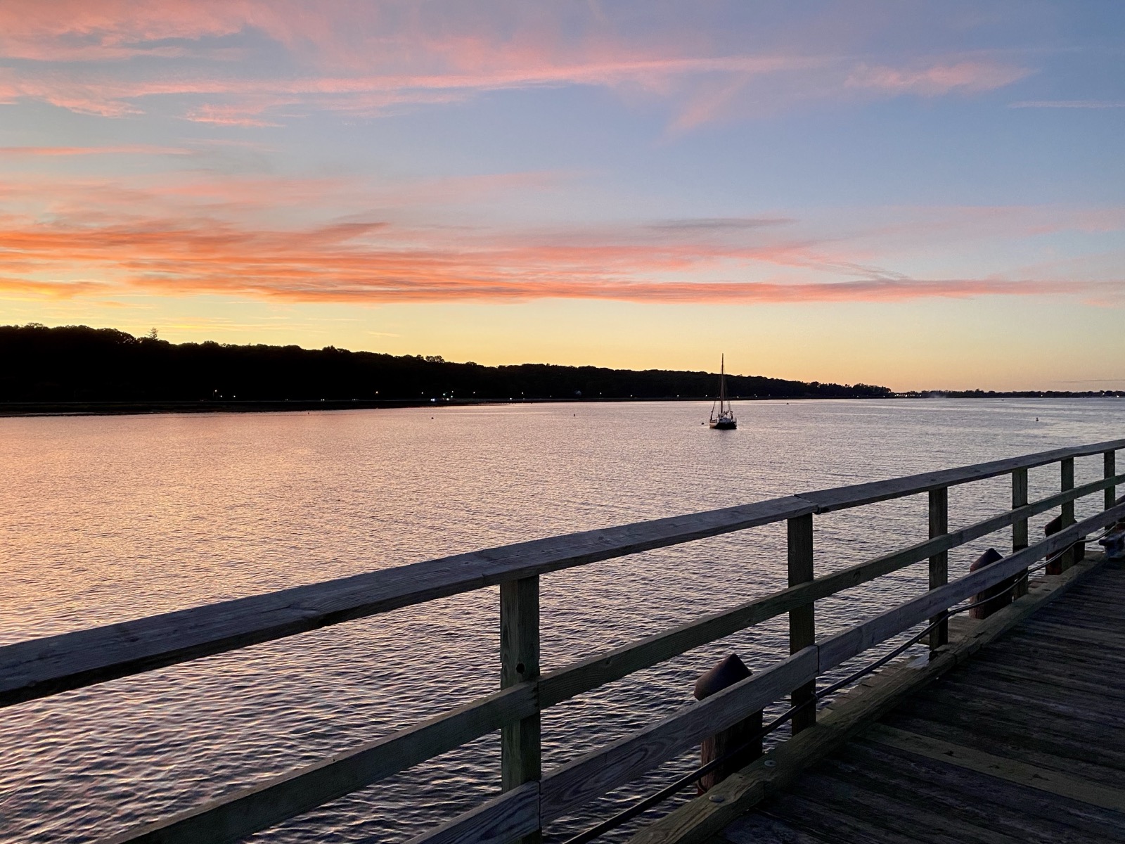 Wooden pier railing over calm bay water at sunset with a sailboat in the distance