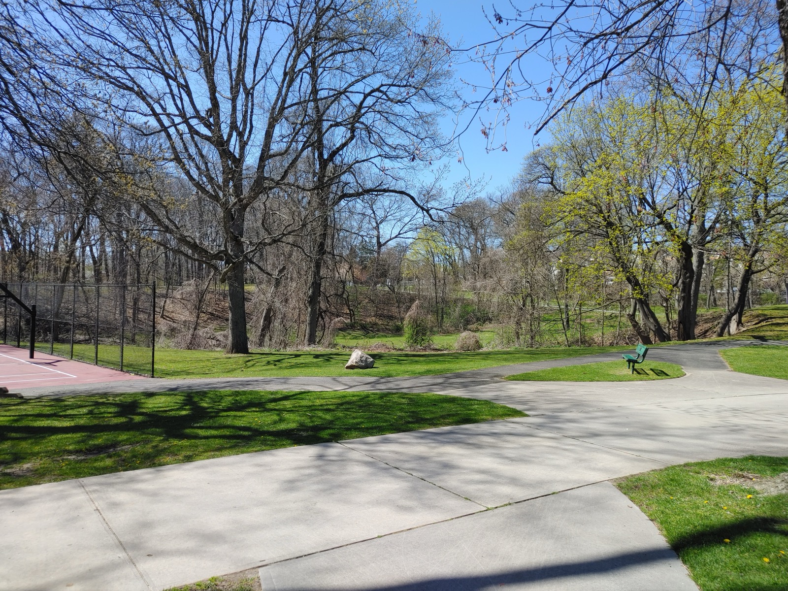Wide paved path curving through Syosset-Woodbury Community Park past bare spring trees and a fenced court